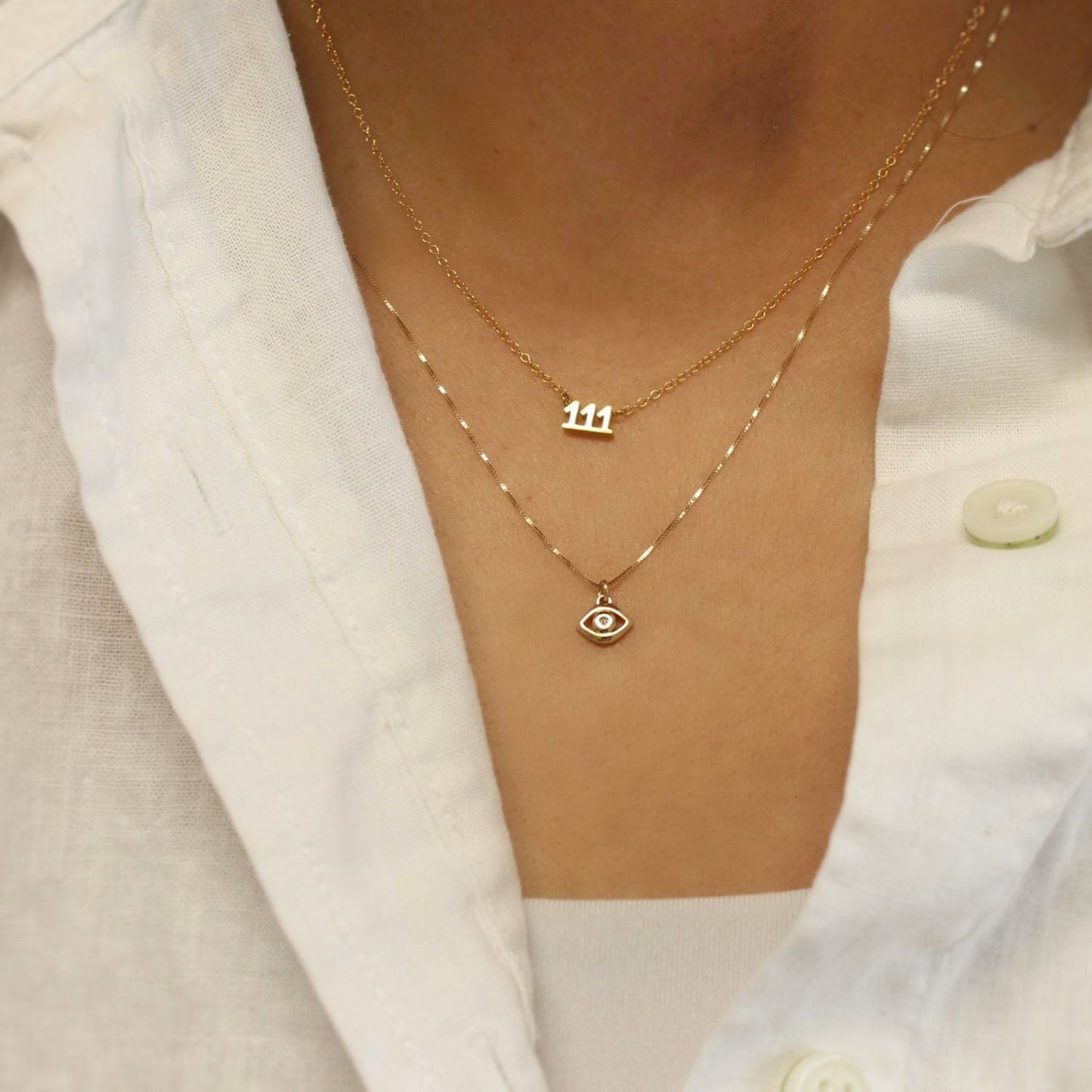 Gold necklaces with number '111' and eye pendant on a white shirt background